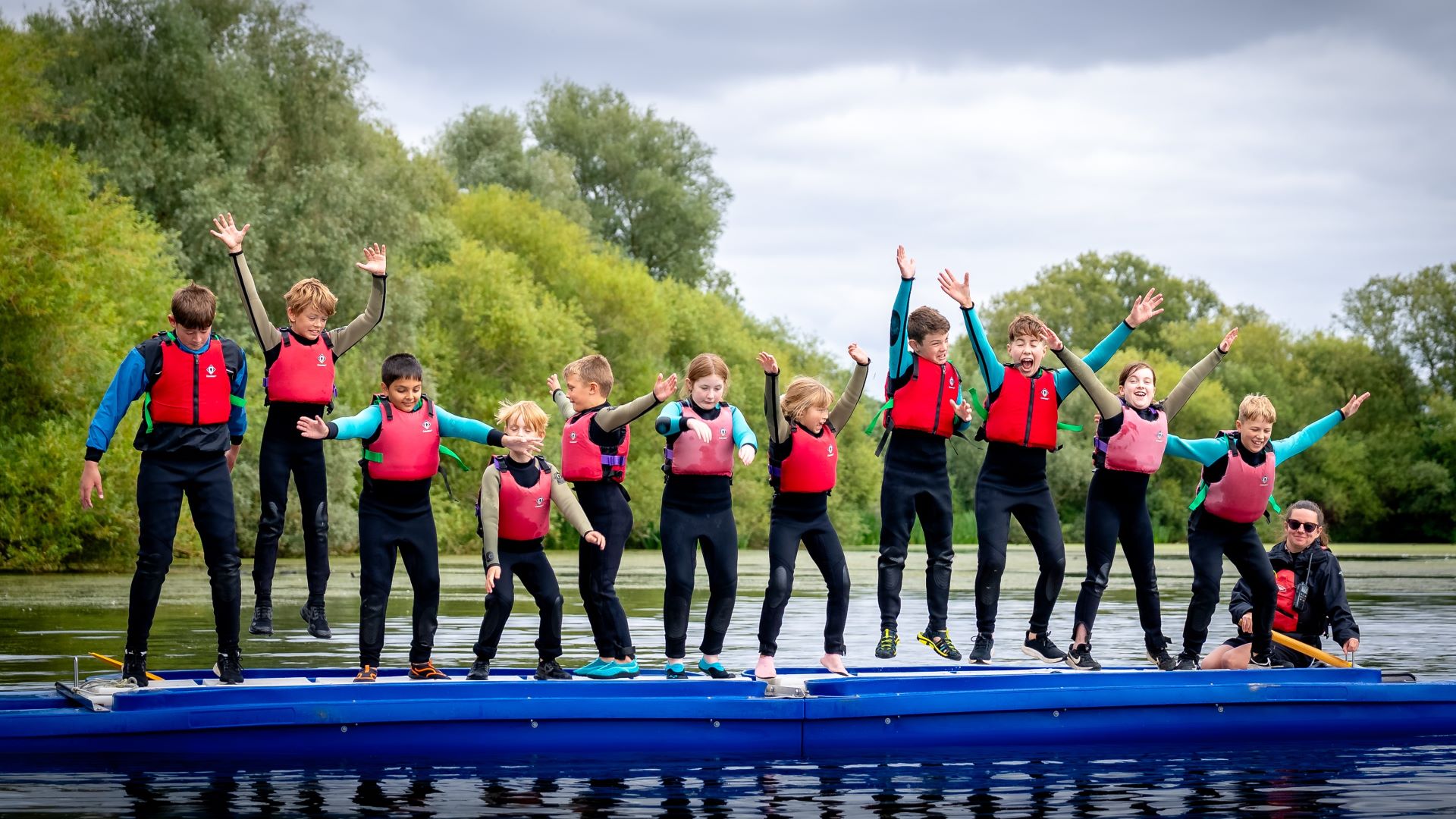 group of school children jumping for joy on a long blue boat in the lake