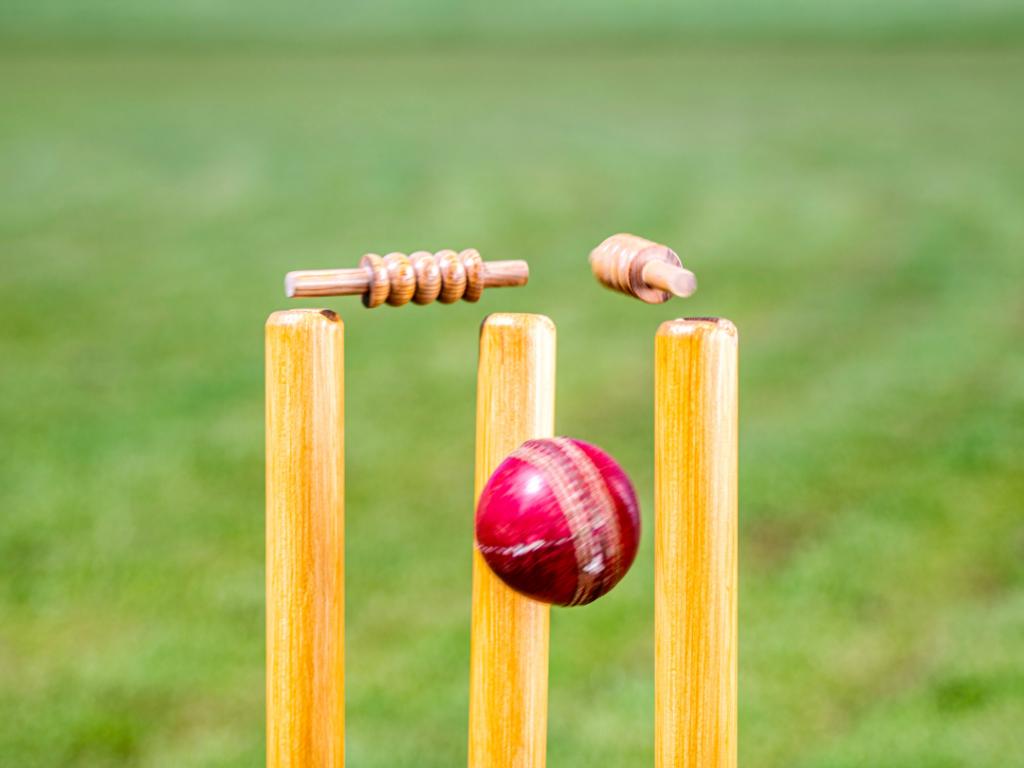 three wooden cricket stumps being hit by a red cricket ball