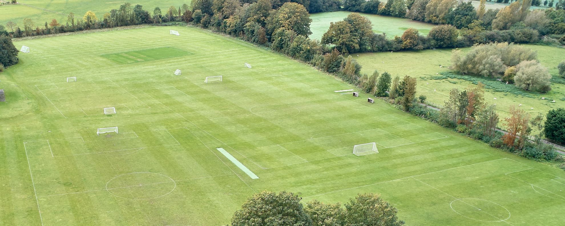 drone image over a large green space which is split into a number of sports pitches with a border of trees surrounding it and countryside beyond