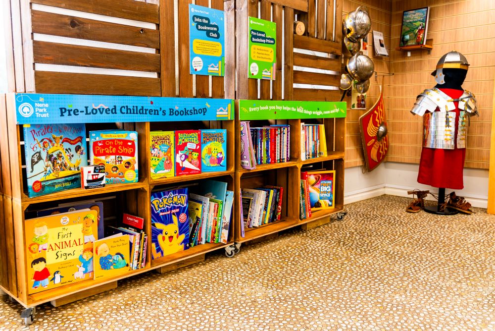 Colourful image of some wooden shelves containing second hand childrens books for sale at the Visitor Centre with a roman costume, helmets and shield in the corner
