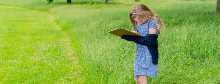 girl in blue school uniform standing in a green grass field with a clipboard