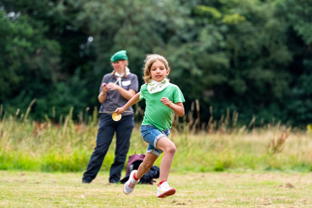 Young girl in a green tshirt and shorts running in a field with a teacher stood behind