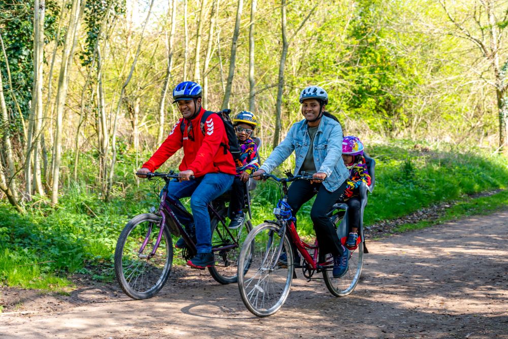 Family with a mum, dad, and two young children cycling in a park on a hard path with trees and grass at the side. The children are in child seats at the back of the parents bikes.