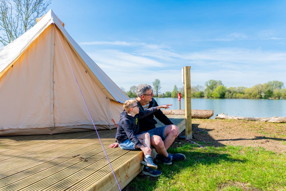 Father and son sitting on a wooden decking next to a large cream coloured bell tent on the side of a lake