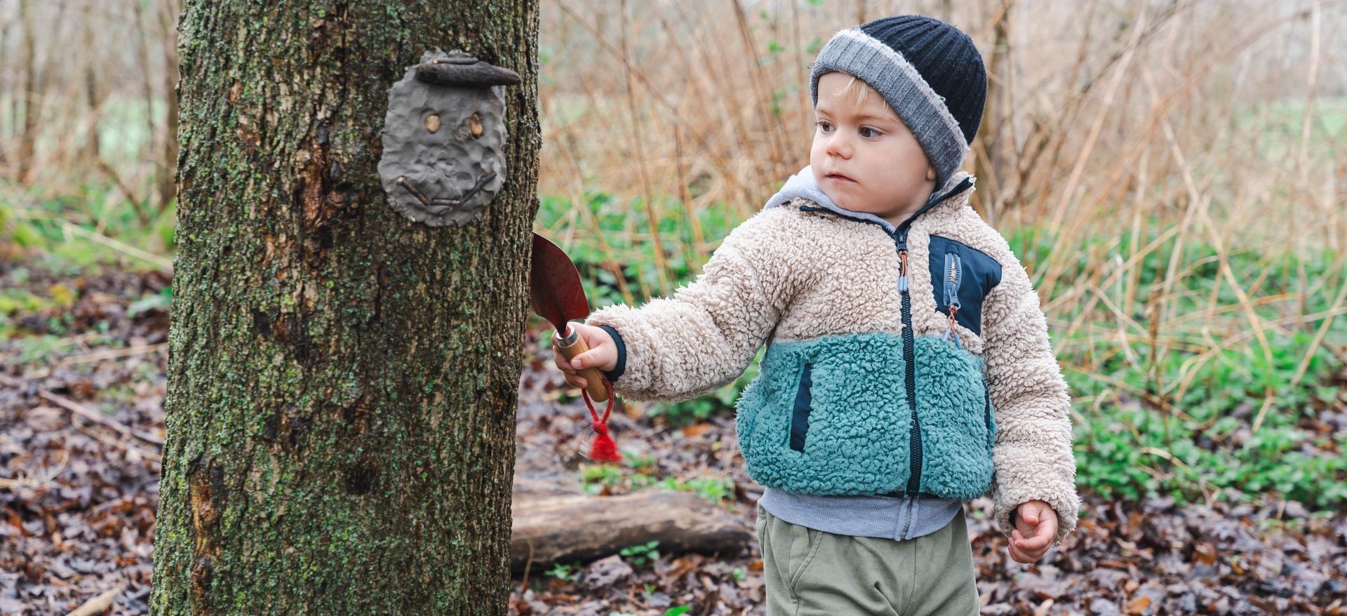 Toddler boy in a fleece jacket and winter hat next to a large tree in the woods