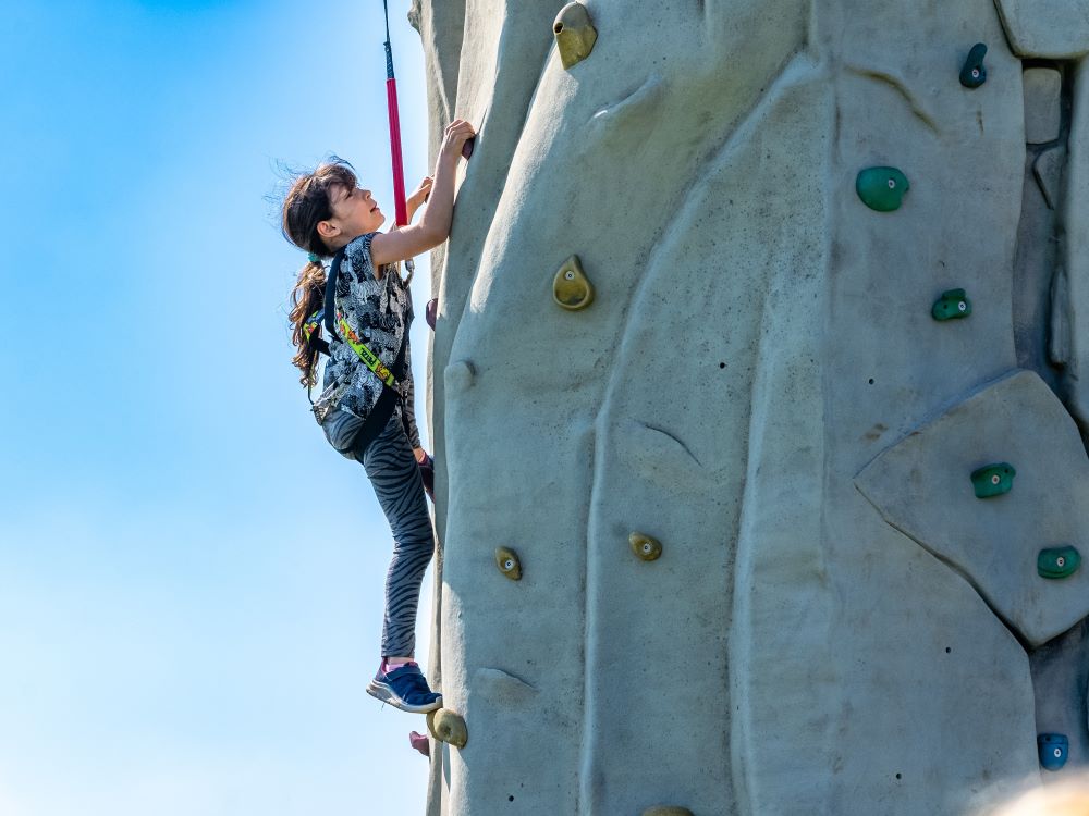 young girl climbing up a grey mobile climbing wall with a blue sky in the background