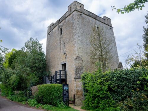 Full view of the impressive stone Longthorpe Tower with wooden staircase to the door and green trees around the edges