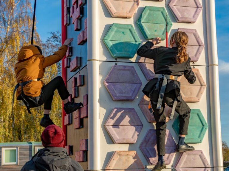 two school children wearing harnesses climbing up a mobile climbing wall