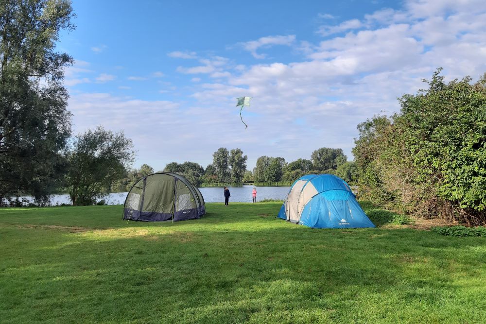 Two pitched tents on a green field next to a large lake surrounded by trees, two children are flying a kite next to the waters edge.
