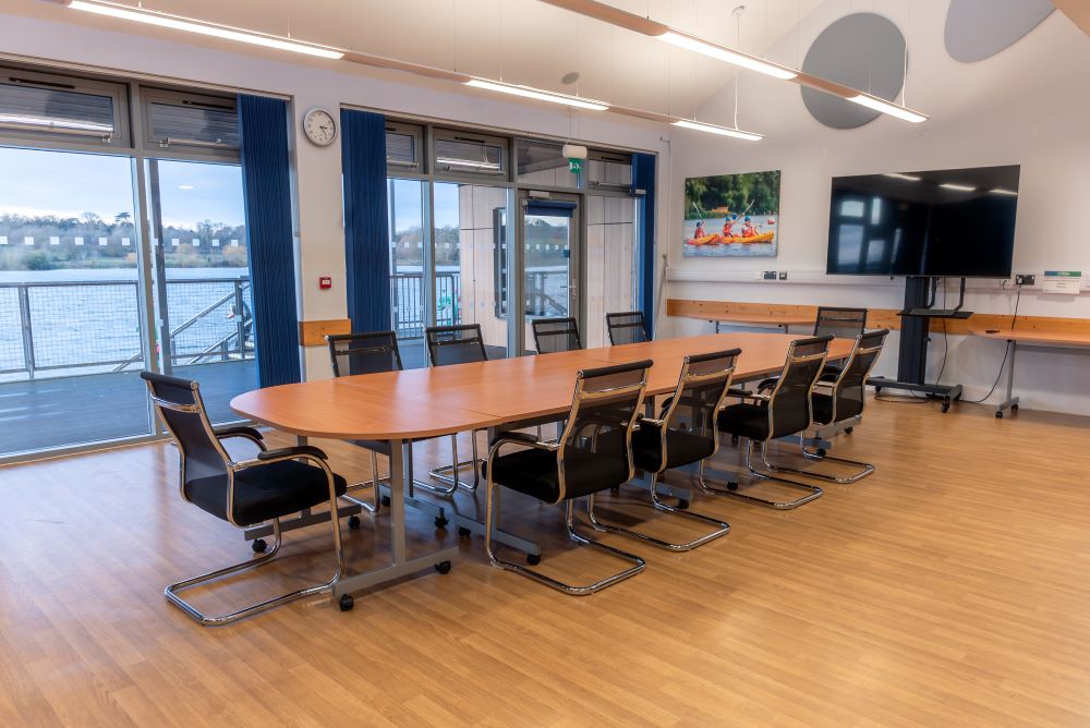 A spacious meeting room with a long wooden table in the middle and 10 black chairs around the outside. A large screen is at the top of the table and floor to ceiling windows show views across a lake.