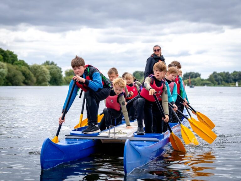 group of school children wearing wetsuits and life jackets paddling on a blue bell boat on a lake
