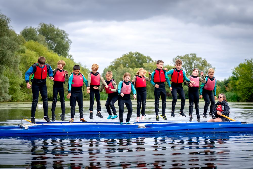 12 children in wetsuits and life jackets in a line jumping for joy on a blue boat sailing on a lake