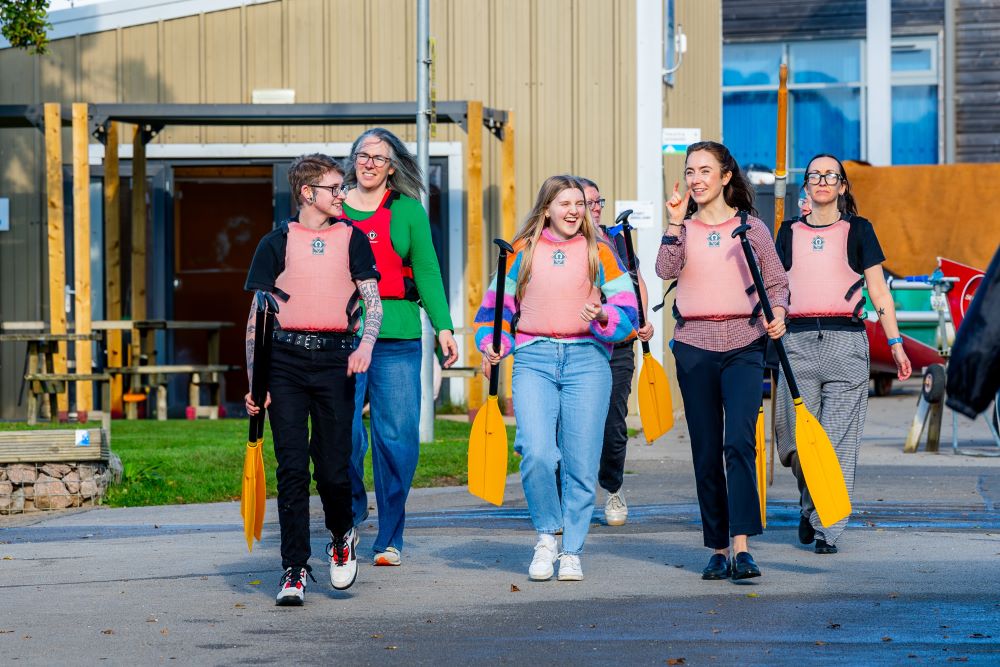 Group of 6 work colleagues heading out to the lake holding yellow paddles and wearing pink life jackets