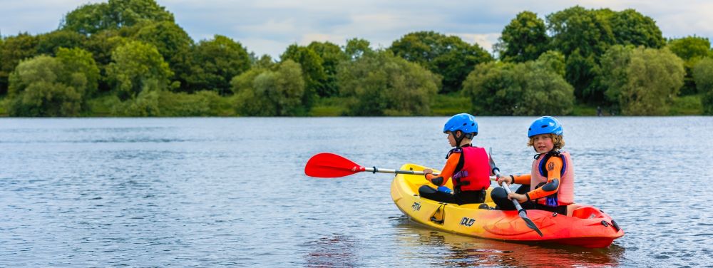 two boys in a red and yellow canoe sailing in a lake with trees in the background