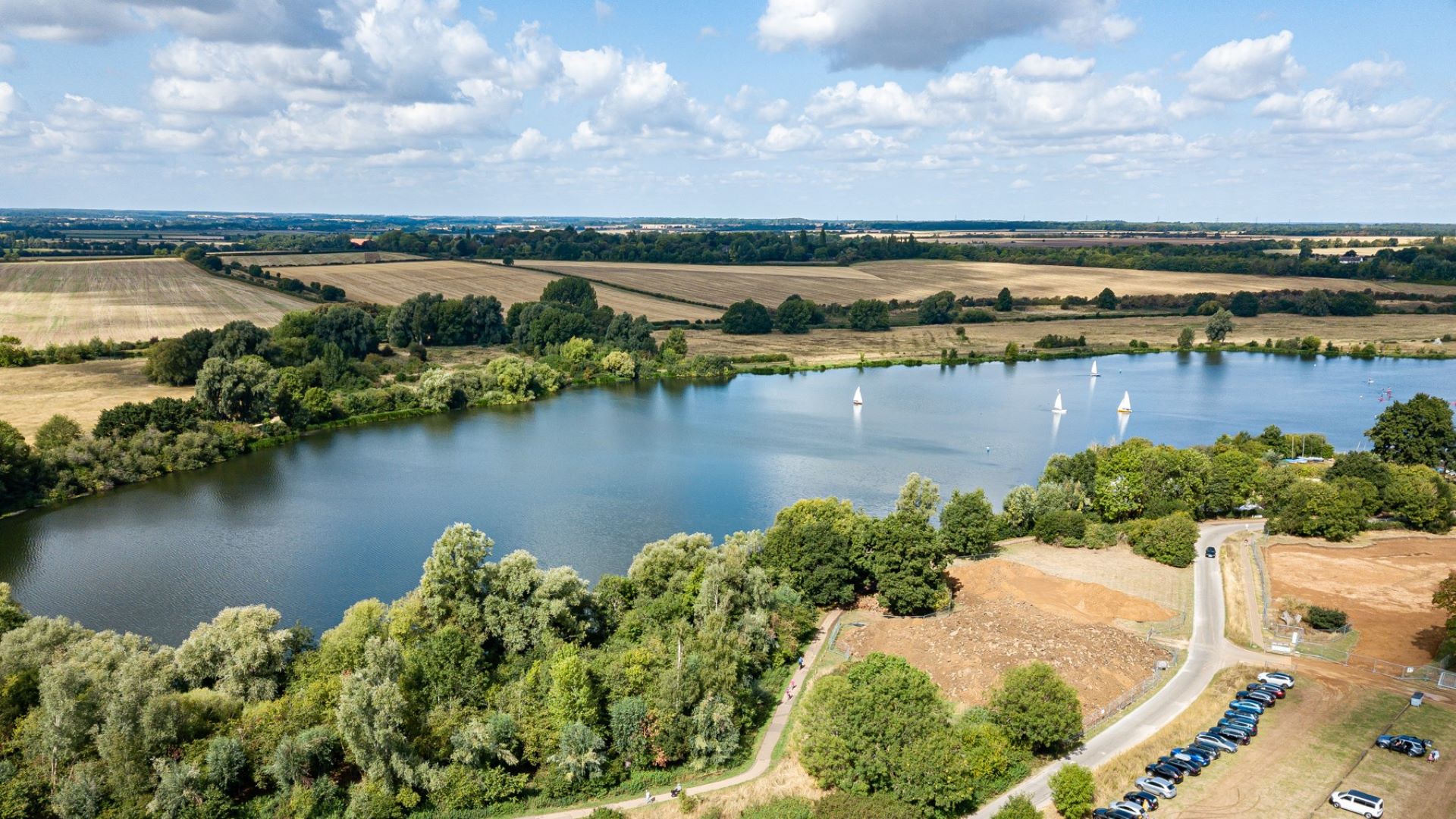 A drone photo showing a large lake surrounded by fields and green trees. On the lake are some sailing boats.