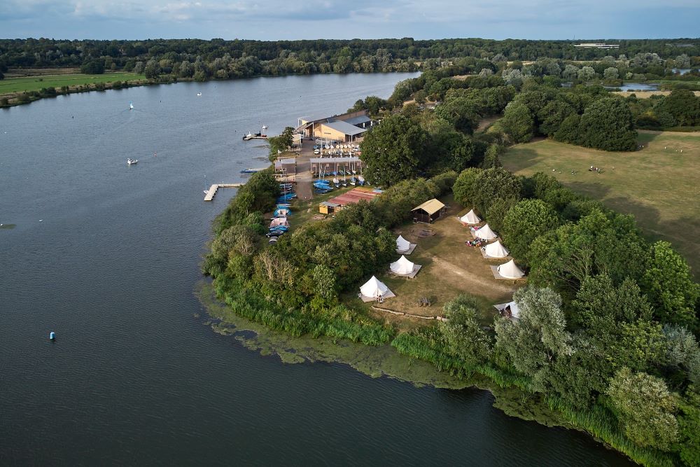 A drone view of a small peaceful campsite of eight white bell tents on the shore of a large lake surrounded by trees and fields and boat yard.