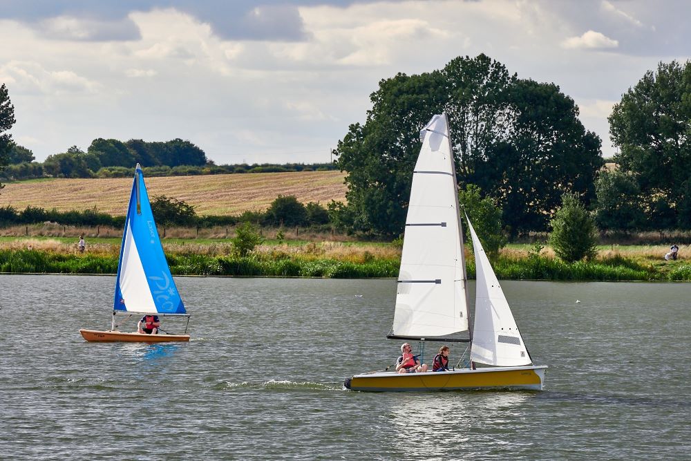 A larger white sail boat and a smaller blue sail boat on a lake with a countryside view