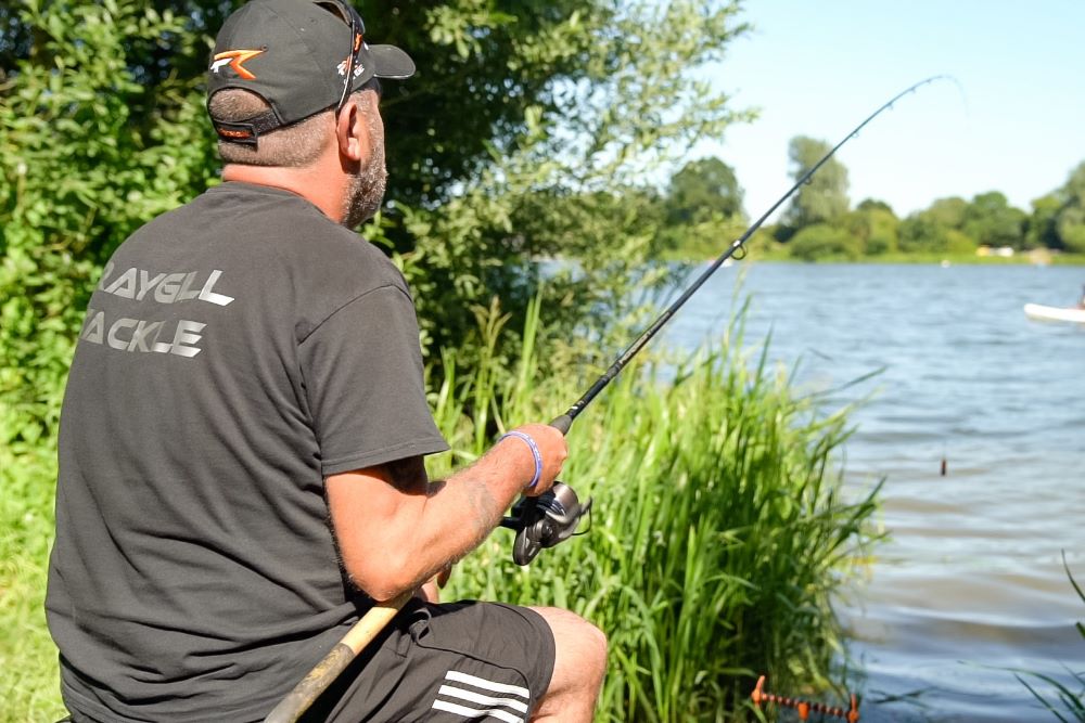 Man in a grey tshirt and shorts fishing on the side of a lake surrounded by green grass and trees