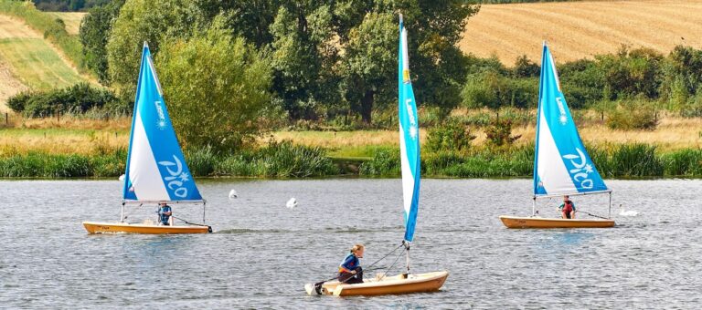 Three small blue sail boats on a lake with fields in the background