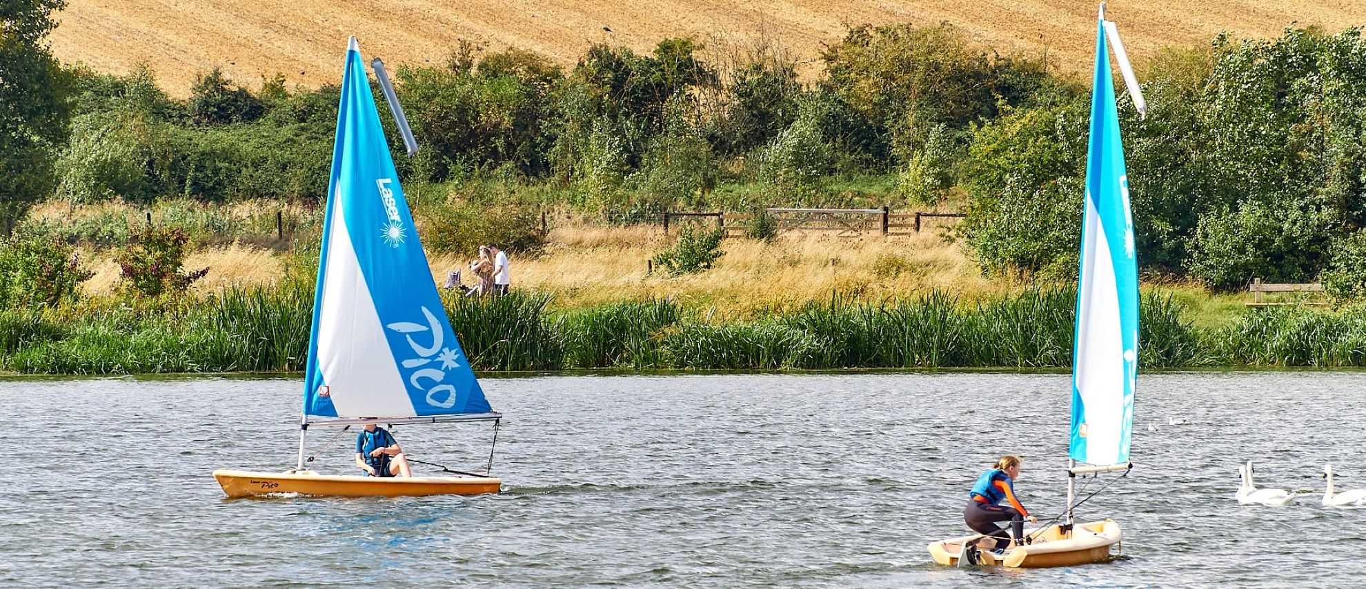 Two small sailing boats with blue sails on a lake with countryside field views