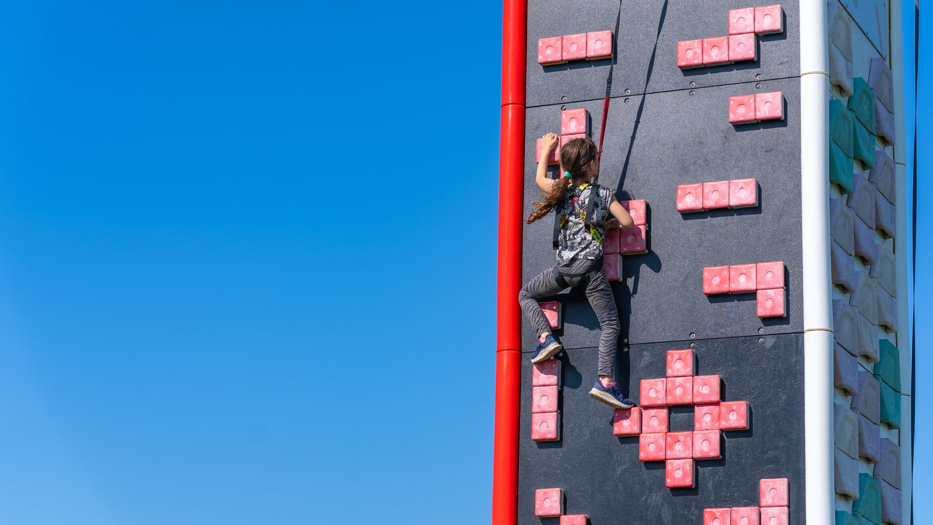 A young girl wearing a harness climbs up a red and black climbing wall. A vibrant blue sky surrounds the climbing wall.