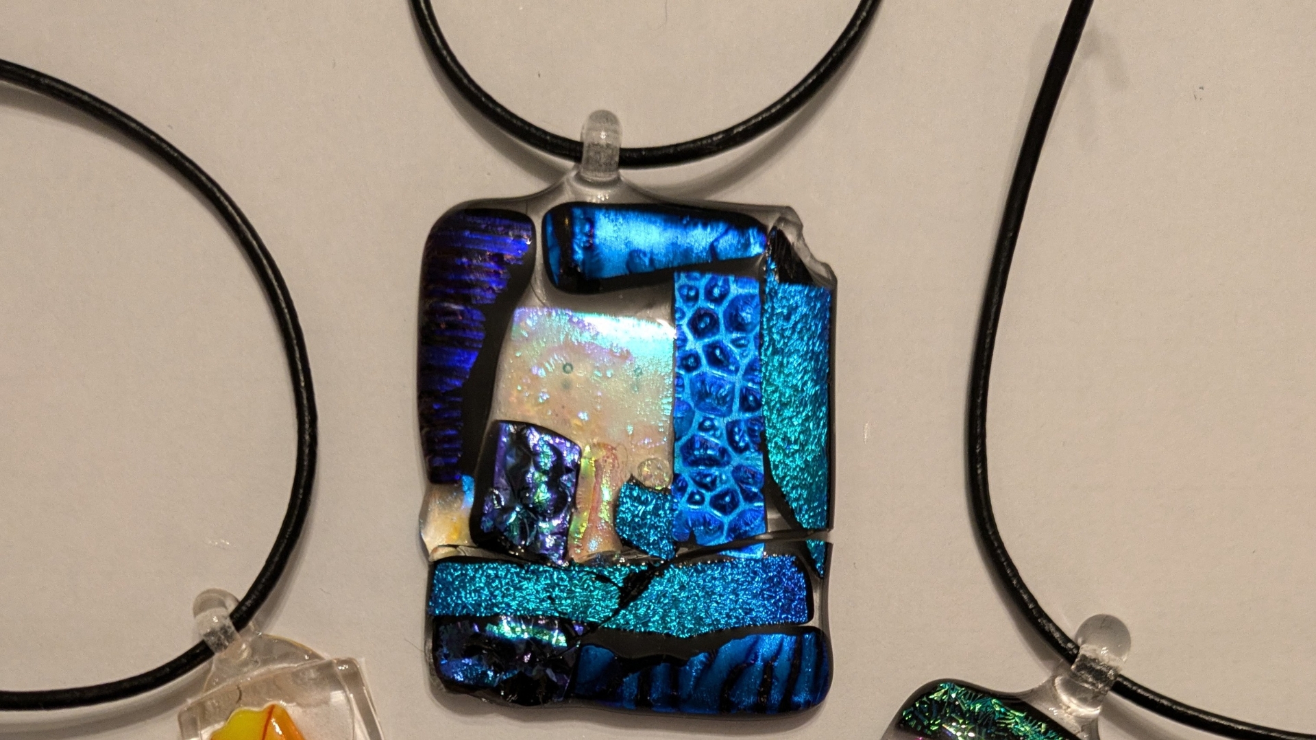 A large blue necklace pendant crafted from different pieces of coloured glass lays on top of a flat, white surface.