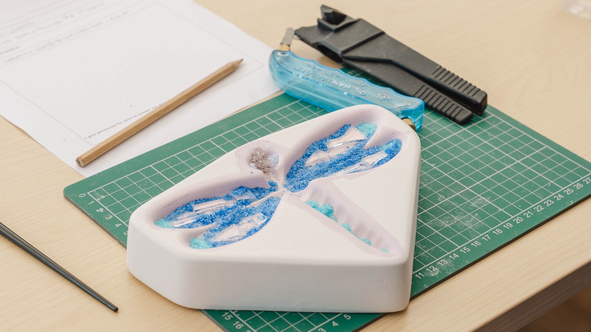 A dragonfly mould sits on a green cutting mat on a flat surface surrounded by several tools including pliers and pencils and paper. Inside the mould are microscopic pieces of blue and white glassful the body of the dragonfly as well as some larger translucent glass shards for the wings