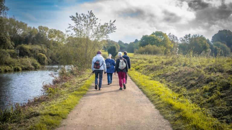 A scenic view of the back of four adults walking along a path with a river on the left and fields to the right