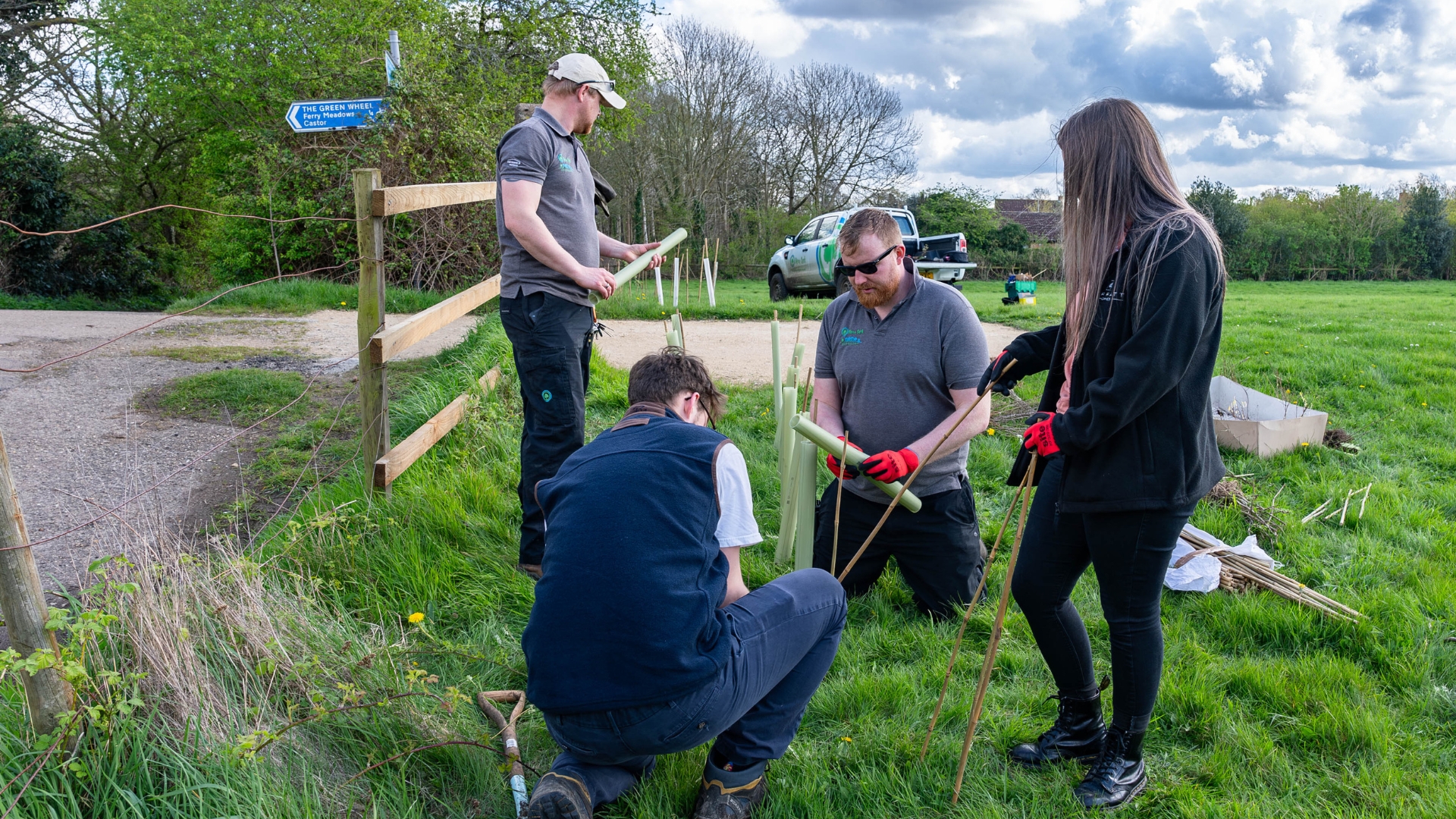 A small group of rangers and volunteers busy themselves planting trees in a green field next to a fenced, public footpath. It is a dry but overcast day.