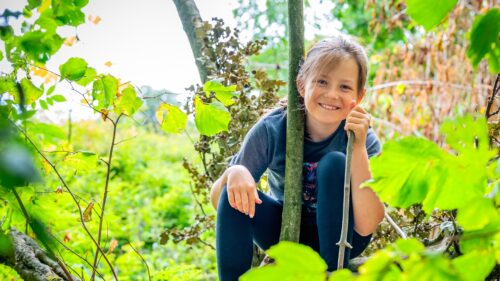 A young girl wearing a t-shirt and leggings sits in a small forest opening, holding a stick. She smiles happily straight at the camera.