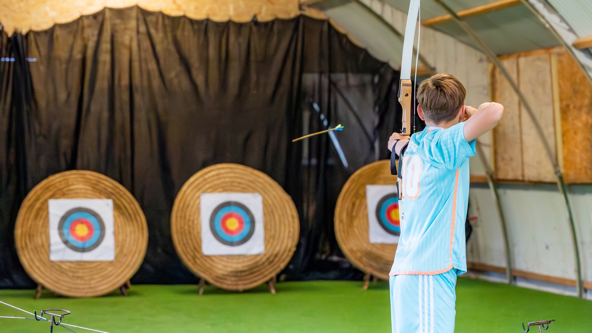 A teenage boy stands indoors with his back to the camera, aiming a bow. He has just released an arrow, which flies off toward 3 visible targets in the background.
