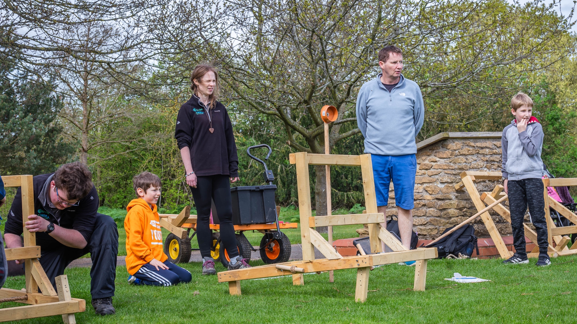 A group of people stand on a small hill looking into the distance. In front of them, a small replica of a Roman catapult, called an onager, has just fired a small soft ball.