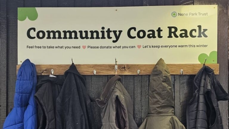 Sign reading Community Coat Rack with a wooden rack beneath, silver hooks with coats on them
