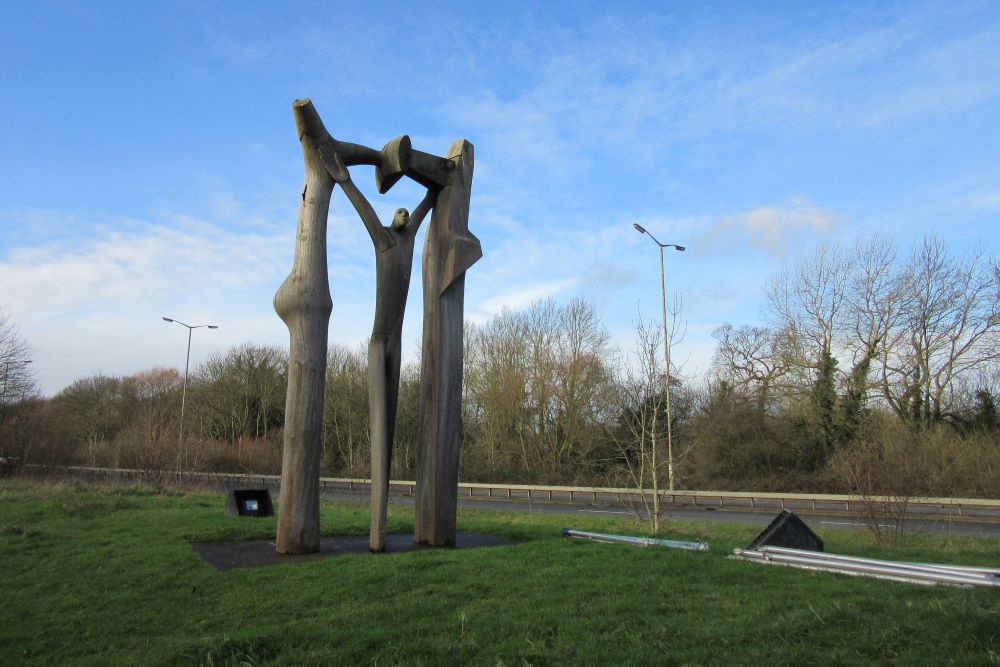 Photo of the Peterborough Arch sculpture in front of a blue sky and road. The sculpture is crafted from wood and depicts a large wooden arch with a person standing inside.