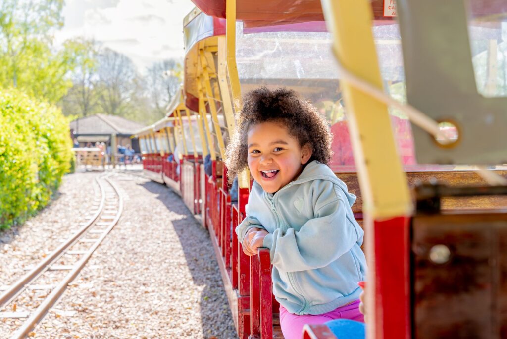 Little girl with brown hair sitting on a red and yellow miniature train smiling at the camera