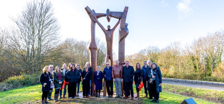Large wooden sculpture of a figure beneath an arch with a group of adults below celebrating it's return