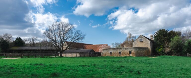 A run down farm site with buildings behind a large green meadow