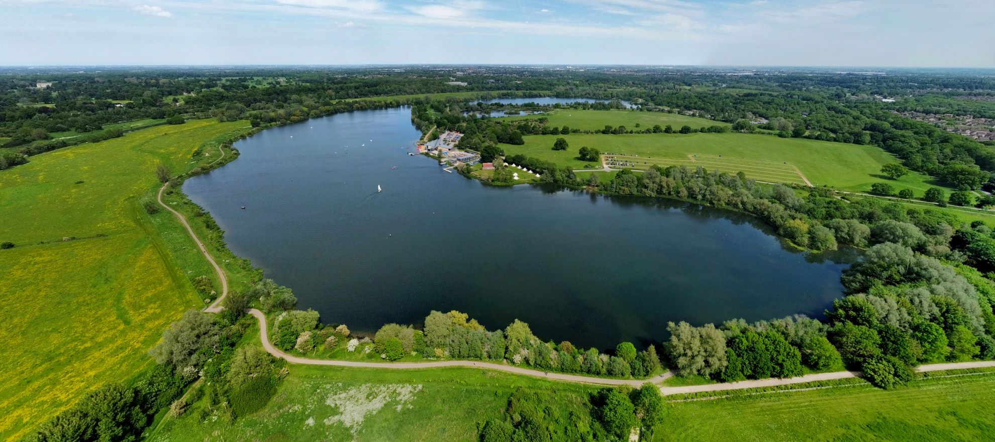 A drone image of a large lake surrounded by green fields, trees and paths