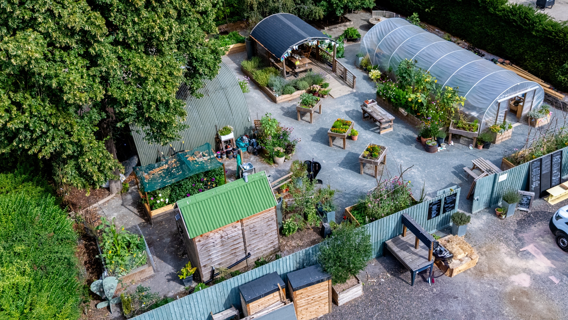An aerial view of an outdoor community plant nursery. The nursery is enclosed by a teal fence and contains a series of raised beds, a polytunnel, seating area under a canopy, potting shed and a composting toilet. The nursery is awash with vibrant colours, it is the middle of summer.
