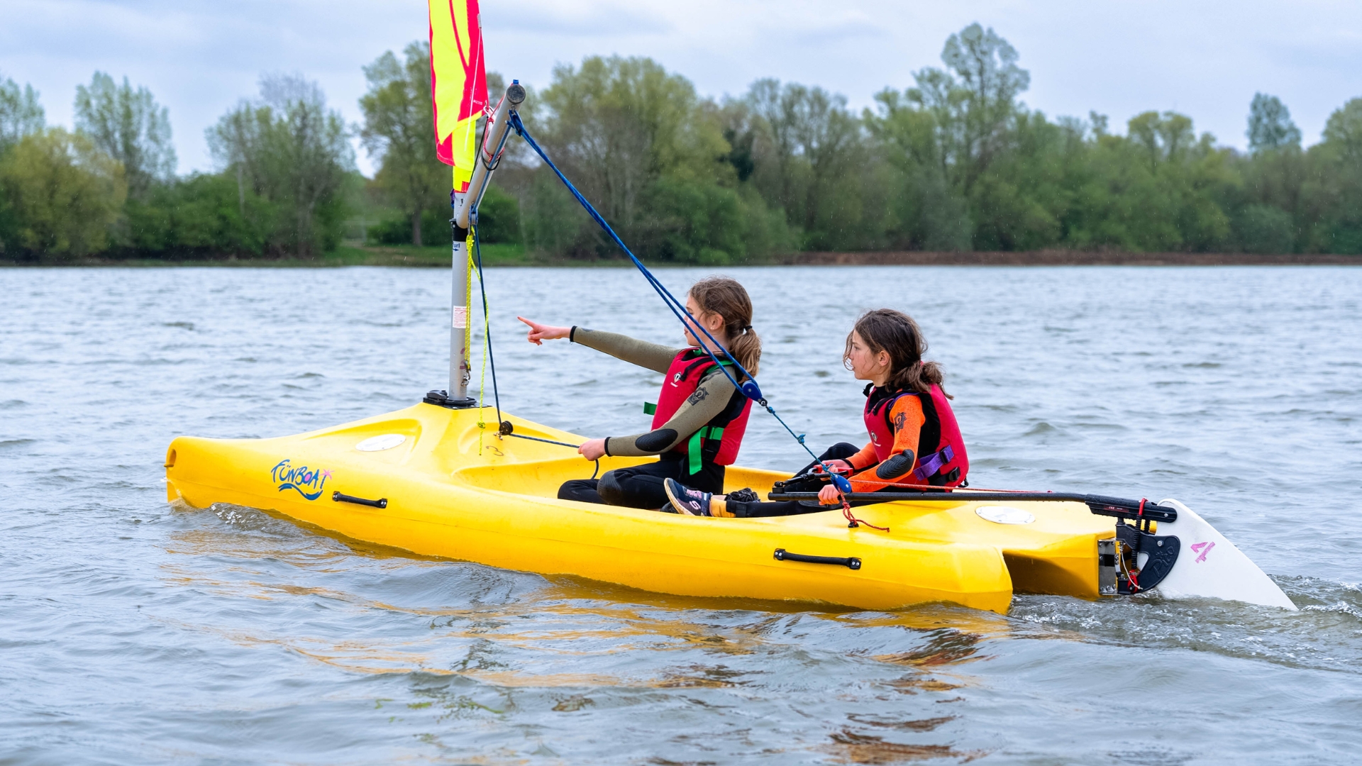 Two young girls sit in a bright yellow sail boat, sailing across the water of a manmade lake. One girl points forward in the direction they intended to sail, the other steers the boat.