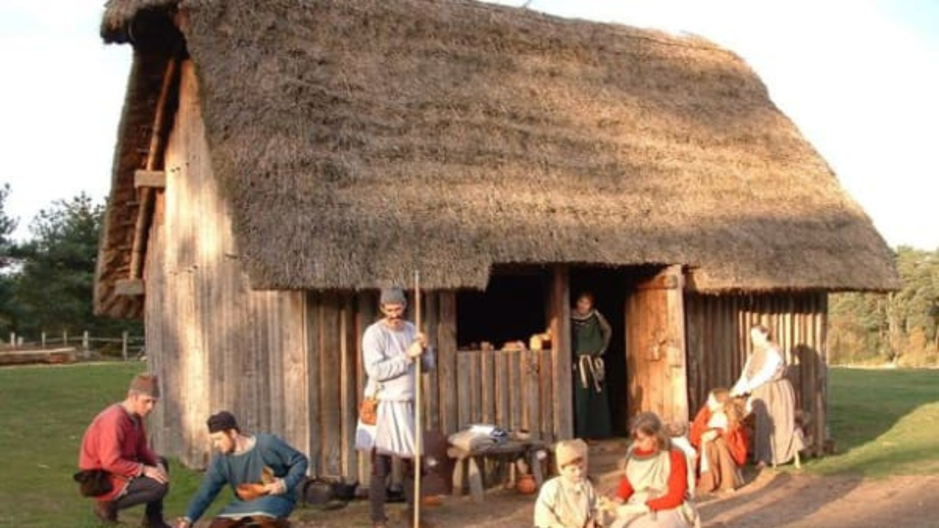 An old Saxon hut with a thatched roof sits on a flat landscape. A Saxon family go about their day in and around the hut, cleaning and preparing food.