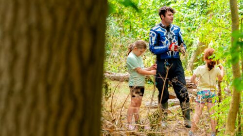 A young, dark-haired male teacher stands in a forest school area talking to two primary school age girls about den-building ideas. A large, out-of-focus tree trunk is to the left of them.