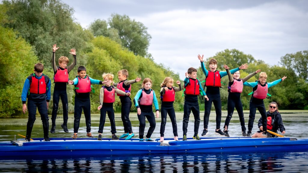 A group of 11 children and teenagers, wearing buoyancy aids, and wetsuits, star jump above a bell-boat that is floating on a large lake. An adult instructor sits at the top of the boat, smiling. The lake is surrounded by vibrant green trees.