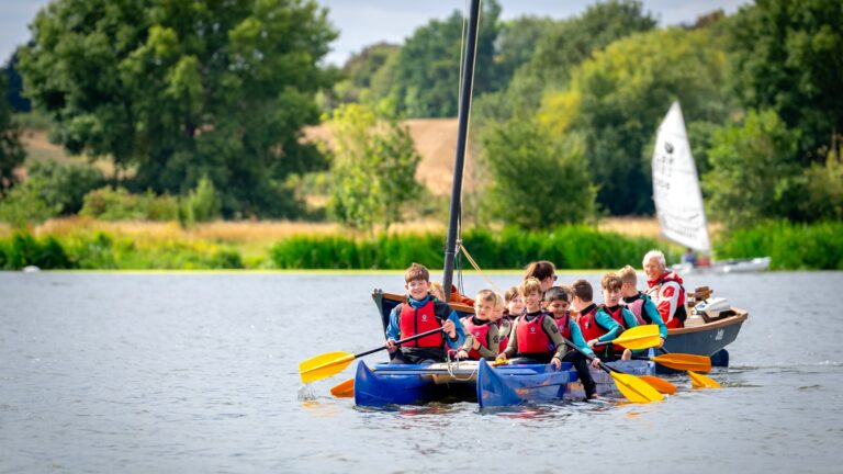 A group of 11 children and teenagers, wearing wetsuits and buoyancy aids, sit in a long blue boat. They hold bright yellow oars as they paddle the boat cooperatively across a large lake. They smile enthusiastically. The lake is surrounded by vibrant green trees and dry, grassy fields.