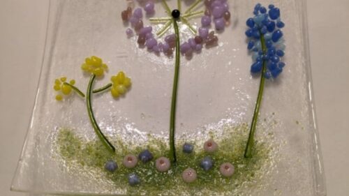 A decorative glass dish sits on a plain, white surface. The dish is made up of transparent glass, with coloured fragments and beads resembling wildflowers in bright, pastel colours.