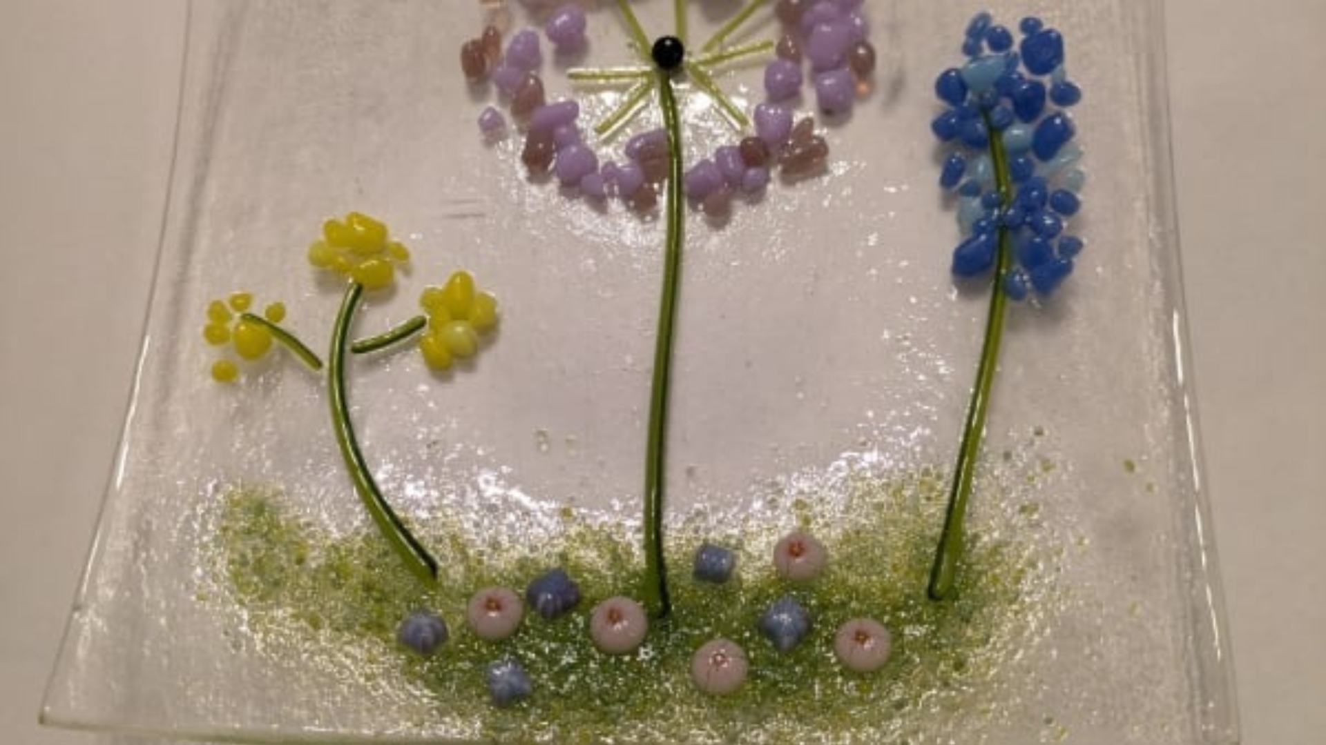 A decorative glass dish sits on a plain, white surface. The dish is made up of transparent glass, with coloured fragments and beads resembling wildflowers in bright, pastel colours.