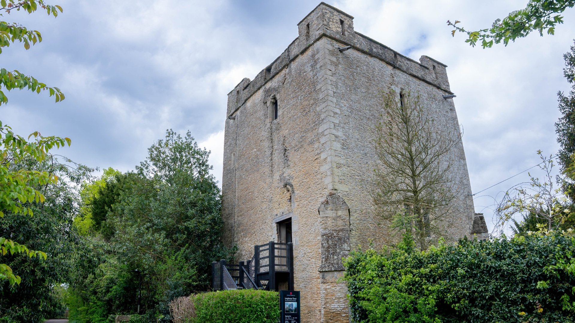 An exterior image looking upward at Longthorpe Tower, a medieval building dating back to the 14th century. It is surrounded by vibrant greenery and a slightly moody sky.
