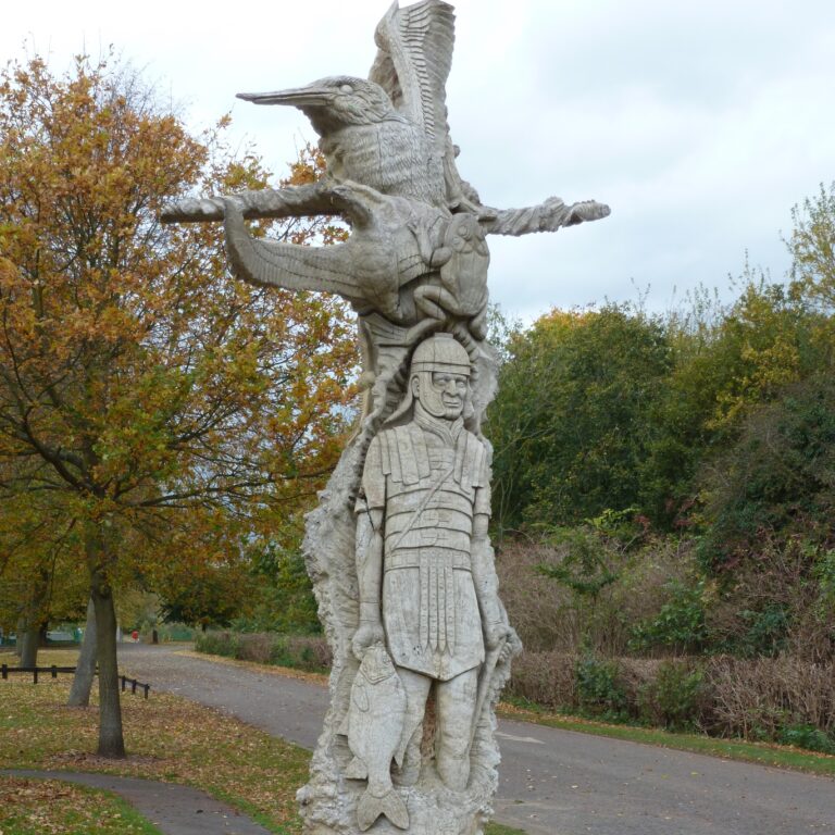 Wooden sculpture of a Roman soldier and park wildlife standing in a park setting.
