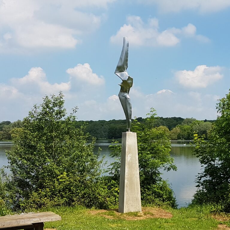 Metal sculpture of a bird in flight standing in front of a lake.
