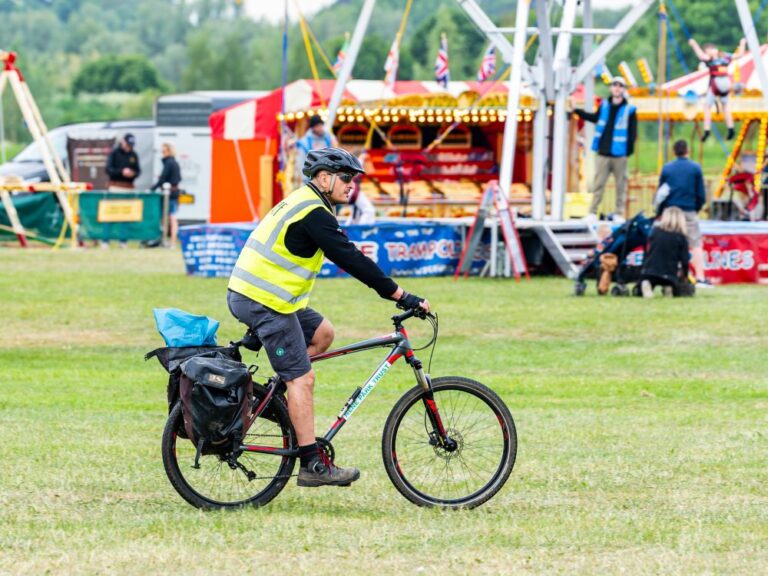 Man on a bike wearing a hi viz jacket. There is a fair ground in the background.